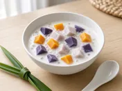 A bowl of bubur cha cha: coconut milk with cubes of orange sweet potato, purple sweet potato and taro and clear sago pearls, a knotted pandan leaf and a spoon beside it