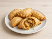 Golden crimped curry puffs on a white plate, one broken open to show the chicken and potato curry filling