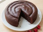 A dense dark chocolate gateau on a white plate with one wedge removed to show the silky crumb, a few raspberries beside it on a pale wooden table