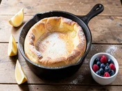 A puffed golden German pancake (Dutch baby) in a cast-iron skillet dusted with powdered sugar, with lemon wedges and mixed berries on a wooden table