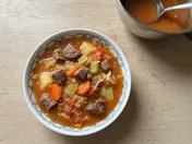 A blue-rimmed white bowl of Hong Kong borscht seen from above: tomato broth with chunks of braised beef, potato, carrot, celery, tomato and cabbage, beside a pot of the soup with a ladle on a pale wooden table