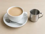 Frothy Ipoh white coffee in a white cup and saucer with a spoon, next to a small stainless steel jug