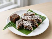 Diamond-shaped pieces of brown palm sugar kuih kosui coated in white grated coconut on a banana leaf, with a block of palm sugar and a pandan leaf