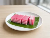 Squares of kuih lapis with neat alternating pink and white steamed layers on a banana leaf, one piece standing on its side to show the layers