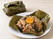 An opened lotus leaf parcel of lo mai gai showing brown glutinous rice with chicken pieces, shiitake mushrooms, dried shrimp and a salted egg yolk, with a wrapped parcel behind