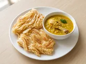 Two flaky golden roti canai on a white plate with a small bowl of chicken curry