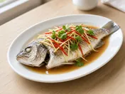 A whole steamed white fish on an oval plate in light soy sauce, topped with shredded ginger, spring onion, red chilli and coriander