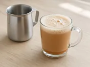 Frothy teh tarik in a glass mug next to a stainless steel pulling jug